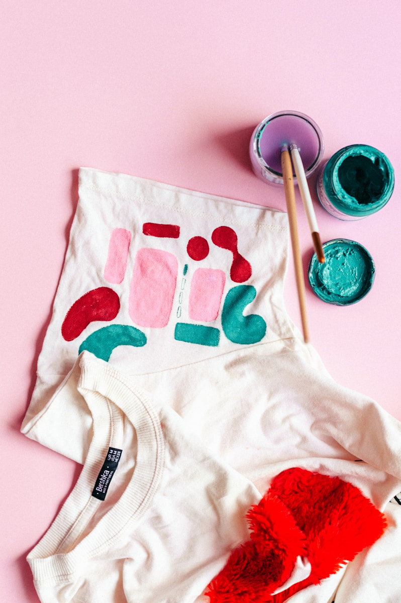 T-shirt blanc et rouge posé à côté de pots de peinture colorée et de pinceaux pour un atelier créatif.